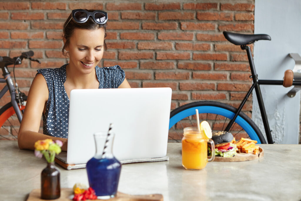 Attractive woman photographer retouching pictures using photo editor, having lunch, sitting in front of generic laptop. Student girl studying online on notebook, smiling joyfully while making progress