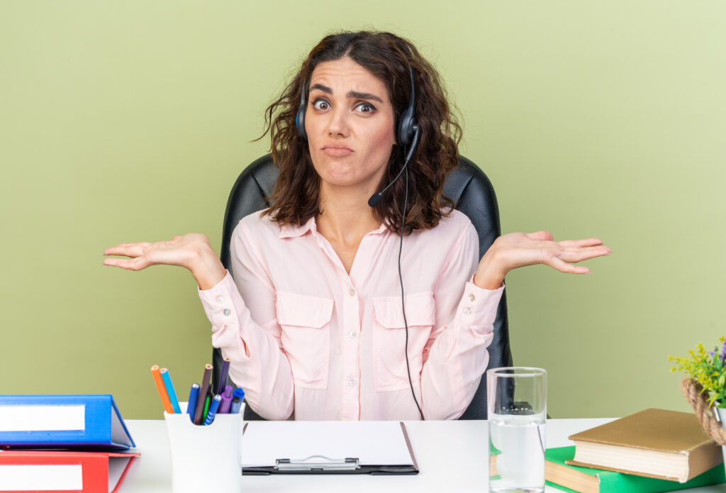 confused pretty caucasian female call center operator on headphones sitting at desk with office tools keeping hands open isolated on green background